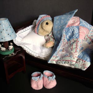 May include: A miniature bedroom scene featuring a teddy bear in a bed with a patchwork quilt and a blue nightcap. A blue lamp with star cutouts sits on a small wooden nightstand. Pink baby booties are placed near the bed.
