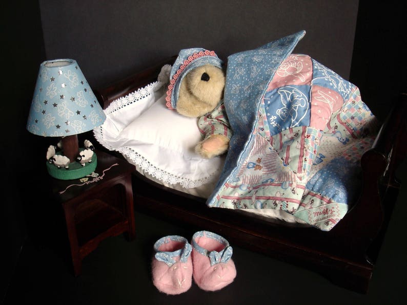 May include: A miniature bedroom scene featuring a teddy bear in a bed with a patchwork quilt and a blue nightcap. A blue lamp with star cutouts sits on a small wooden nightstand. Pink baby booties are placed near the bed.