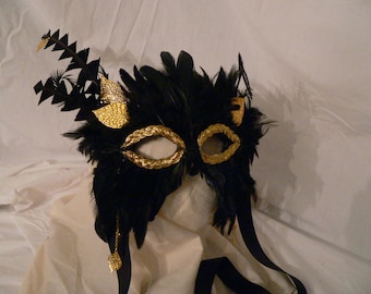 Puede incluir: Una máscara de carnaval negra con detalles dorados y plumas. La máscara tiene un borde dorado alrededor de los agujeros de los ojos y un diseño de plumas doradas en el lateral.