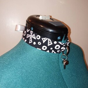 Pode incluir: Um colar choker preto e branco com um pingente de sino de prata. O colar está em um manequim turquesa.