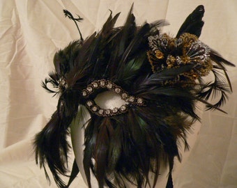 Puede incluir: Una máscara de carnaval negra con plumas y pedrería. La máscara está decorada con detalles dorados y plateados.