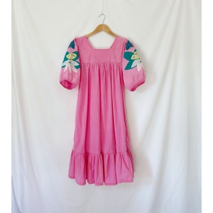 K&ouml;nnte beinhalten: Ein rosafarbenes Midikleid mit quadratischem Ausschnitt und kurzen Puff&auml;rmeln. Die &Auml;rmel sind mit floralen Stickereien in Gr&uuml;n-, Wei&szlig;- und Gelbt&ouml;nen verziert.