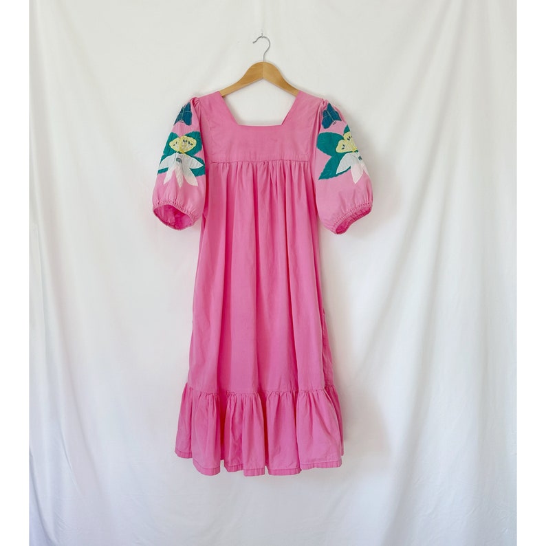 K&ouml;nnte beinhalten: Ein rosafarbenes Midikleid mit quadratischem Ausschnitt und kurzen Puff&auml;rmeln. Die &Auml;rmel sind mit floralen Stickereien in Gr&uuml;n-, Wei&szlig;- und Gelbt&ouml;nen verziert.
