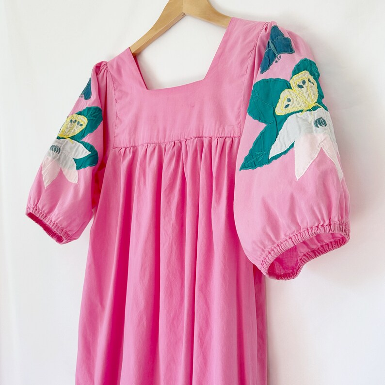 K&ouml;nnte beinhalten: Ein rosa Kleid mit quadratischem Ausschnitt und Puff&auml;rmeln. Die &Auml;rmel sind mit gr&uuml;nen Bl&auml;ttern und wei&szlig;en Blumen bestickt.