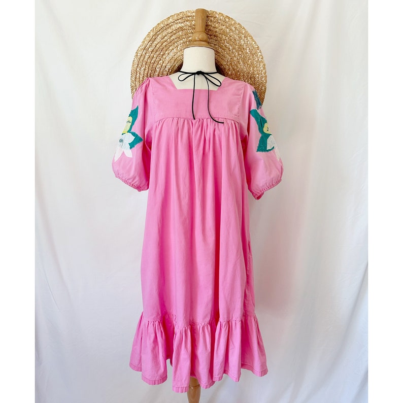 K&ouml;nnte beinhalten: Ein rosa Baumwollkleid mit quadratischem Ausschnitt und gestickten Blumen an den &Auml;rmeln. Das Kleid hat einen R&uuml;schen-Saum und wird mit einem Strohhut getragen.
