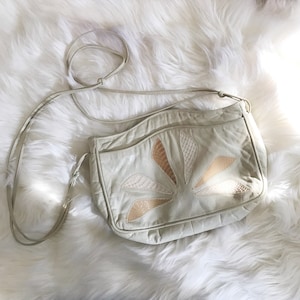 May include: A white leather shoulder bag with a floral design made of beige and brown leather. The bag has a long strap.