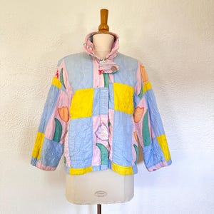 May include: A quilted jacket in a patchwork design, featuring squares of light blue, yellow, and pink fabric. The jacket has a high collar and is adorned with floral appliqués. The jacket is displayed on a mannequin.