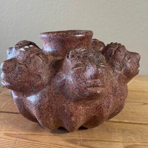 May include: A red clay pot with four faces sculpted into the sides. The pot has a speckled texture and a round opening at the top.