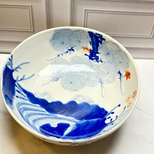 May include: A white ceramic bowl featuring hand-painted blue wave patterns along the bottom, light blue floral motifs, and small orange accents. The interior surface displays a glossy finish with detailed brushwork depicting water and flowers.