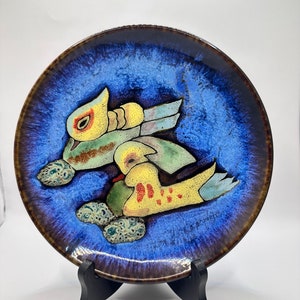 May include: A decorative ceramic plate with a blue and brown glazed finish. The plate features a design of stylized birds in yellow, green, and blue. The plate is displayed on a black stand.