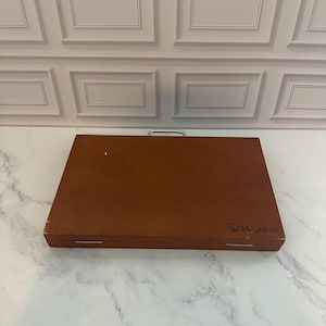 May include: A rectangular wooden carrying case with a smooth brown finish, featuring a silver metal handle on the top edge, two silver hinges on the front, and the word "MIRADOR" printed in black on the lower right corner of the lid.