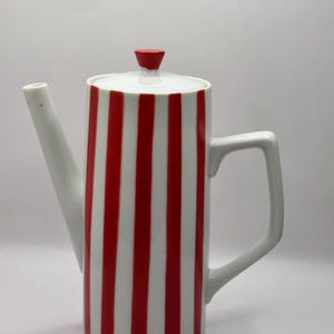 May include: A white ceramic coffee pot with vertical red stripes. The pot has a long spout, a handle, and a lid with a red knob. The design is simple and modern, suitable for serving coffee or tea.