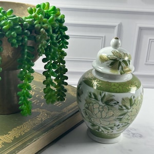 May include: A white porcelain ginger jar featuring a green and gold floral pattern with a matching lid. The jar has a rounded body and a tapered base, decorated with intricate botanical illustrations and gold filigree accents on the neck.