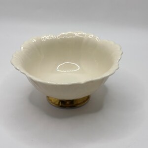 May include: A cream-colored, scalloped-edge bowl with a gold-colored base. The bowl has a rounded shape and sits on a short, pedestal base. The interior of the bowl is smooth and reflective. The bowl is likely made of ceramic or porcelain.
