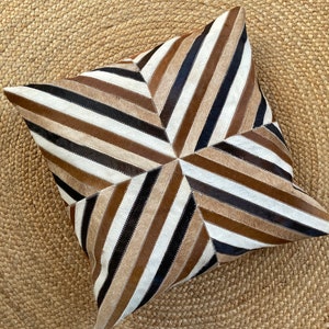 May include: A square throw cushion featuring a geometric starburst pattern composed of alternating diagonal strips of hair-on-hide leather in shades of dark brown, light brown, tan, and white, meeting at the centre of the cushion surface.