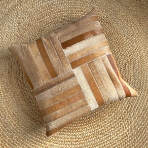 May include: A square throw pillow featuring a patchwork design of cowhide strips in varying shades of tan, beige, cream, and dark brown. The strips are arranged in a geometric pattern with alternating horizontal and vertical orientations.