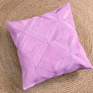 May include: A square, lilac-coloured decorative cushion with a geometric pattern. The cushion is made of a soft material and features a stitched design. The cushion is placed on a woven, tan-coloured rug.