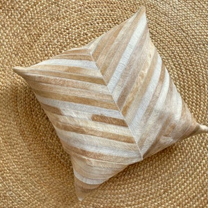Peut inclure: Un coussin carré décoratif avec un motif en chevrons. Le coussin est fait de peau de vache beige et crème. Le coussin est posé sur un tapis en jute tissé.