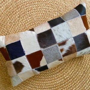 May include: A rectangular throw cushion constructed from a patchwork of cowhide squares. The cushion features a multi-coloured checkerboard pattern in shades of brown, black, white, grey, and tan, with visible stitching connecting each individual hide square.