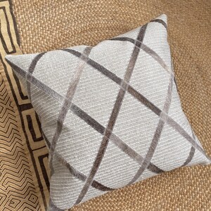 May include: A square throw cushion featuring a light grey textured fabric base with a prominent dark grey and brown crisscross grid pattern. The grid lines appear to have a soft, hair-on-hide texture, contrasting with the ribbed weave of the cushion.