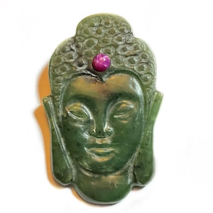 May include: A carved green jade Buddha pendant with a pink gemstone on the forehead. The Buddha's face is serene, with closed eyes and a gentle smile. The pendant is a detailed carving of the Buddha's head.
