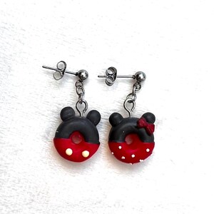 Puede incluir: Un par de pendientes colgantes con dijes en forma de dona de color negro y rojo con lunares blancos. Un dije tiene un lazo negro y el otro tiene orejas de Mickey Mouse.