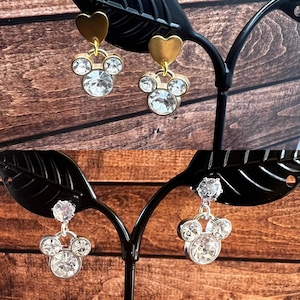 May include: Two pairs of dangle earrings displayed on a black stand. The top pair features gold-tone heart studs with clear crystal Mickey Mouse-shaped charms. The bottom pair features clear crystal studs with clear crystal Mickey Mouse-shaped charms.