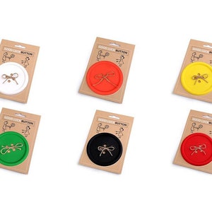 Könnte beinhalten: Eine Sammlung von sechs farbenfrohen "Good Luck Button"-Neuheiten. Jeder Knopf hat eine andere Farbe: Weiß, Orange, Gelb, Grün, Schwarz und Rot. Die Knöpfe sind auf einer Kartonrückseite mit einem Schnur-Schleifen-Design verpackt.