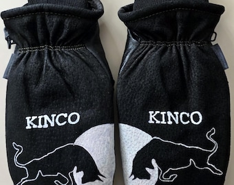 Puede incluir: Un par de manoplas de cuero negro con puños acanalados negros. Cada manopla presenta el texto blanco KINCO y un gráfico blanco de la silueta de un toro sobre un fondo circular blanco. El texto AMT Designs es visible en la esquina superior.