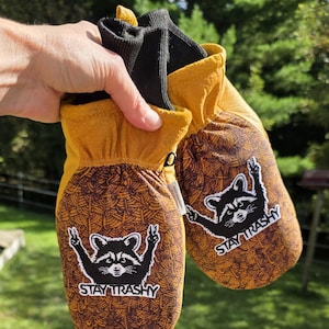May include: A pair of mustard yellow and brown patterned mittens with black ribbed cuffs. The mittens feature a graphic of a black raccoon making a peace sign with both paws and the white text STAY TRASHY printed below the raccoon graphic.