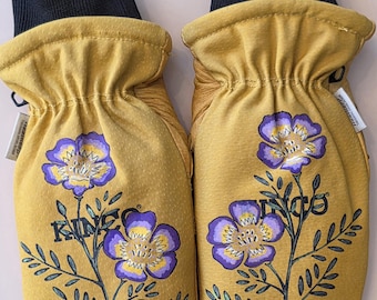 May include: A pair of mustard yellow mittens with a black cuff and a floral design. The floral design features purple and yellow flowers with green stems and leaves. The word "KING" is printed on the mittens. The text "AMT Designs" is in the upper right corner.