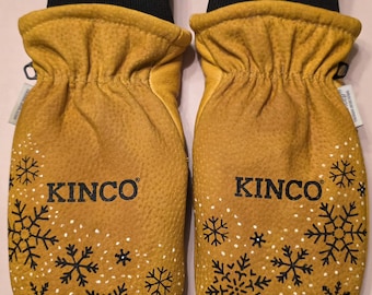 May include: A pair of mustard yellow leather mittens with black snowflake designs and the brand name "KINCO" printed on each mitten. The cuffs are black ribbed knit, and the mittens have a small yellow leather tab. The text "AMT Designs" is visible.