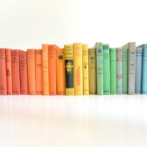 May include: A collection of vintage books arranged in a rainbow gradient. The spines transition from red to orange, yellow, green, and blue. The books are displayed on a white surface against a white background.