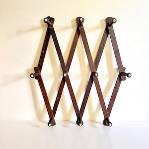 May include: An expandable dark brown wooden wall rack featuring a lattice design with ten pegs. Each peg is tipped with a small white circular accent. The accordion-style structure allows for adjustable width, creating a diamond-shaped pattern.