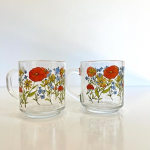 May include: Two clear glass mugs with handles, decorated with a floral pattern. The design features orange poppies, yellow daisies, and blue wildflowers with green stems and leaves. The mugs are set against a white background.