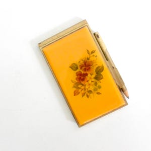 May include: Vintage compact with a bright orange surface and a floral design in shades of red, brown, and green. The compact has a gold-colored frame and a pencil is attached to the side.