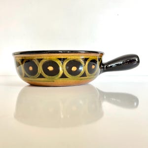 May include: A ceramic bowl with a handle, featuring a black rim and handle. The bowl's exterior is adorned with a repeating pattern of black circles outlined in green and accented with gold dots. The bowl is likely used for serving or cooking.