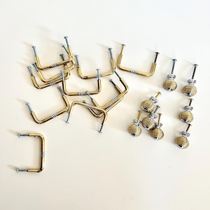 May include: A collection of gold-toned cabinet pulls and knobs. The pulls are U-shaped with a ribbed center, and the knobs have a ribbed, spherical design. Each piece includes silver-colored screws for installation. The hardware is arranged on a white surface.