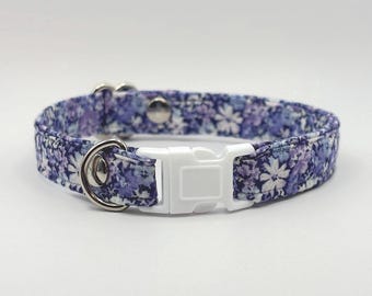 May include: A floral dog collar with a white plastic buckle and silver-tone metal hardware. The collar features a pattern of small white flowers on a blue and purple background. The collar is approximately 1 inch wide.