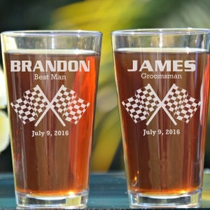 May include: Two clear pint glasses filled with a brown liquid. Each glass is etched with a name, role, checkered flags, and the date July 9, 2016. The glass on the left says "BRANDON Best Man". The glass on the right says "JAMES Groomsman".
