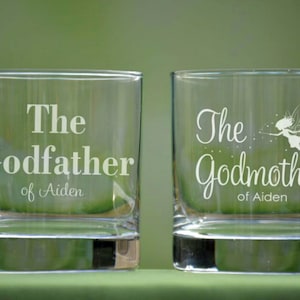 May include: Two clear square-shaped glasses with white text. One glass reads "The Godfather of Aiden", the other reads "The Godmother of Aiden" with a fairy illustration. The glasses are on a green surface.