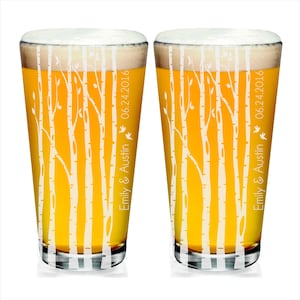 May include: Two pint glasses filled with beer and foam. The glasses are decorated with a white birch tree design. The text "Emily & Austin" and "06.24.2016" are printed on the side of each glass, along with a small bird graphic.