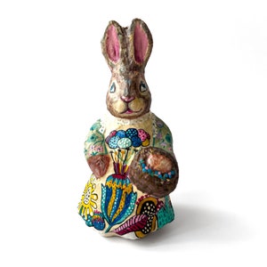 May include: A hand-painted paper mache bunny figurine with brown fur, pink ears, and a colorful floral dress. The bunny is holding a basket of eggs.
