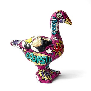 May include: A pink, paper mache goose sculpture with a comic book design. The goose is covered in colorful words like "cool", "oops", and "wow".