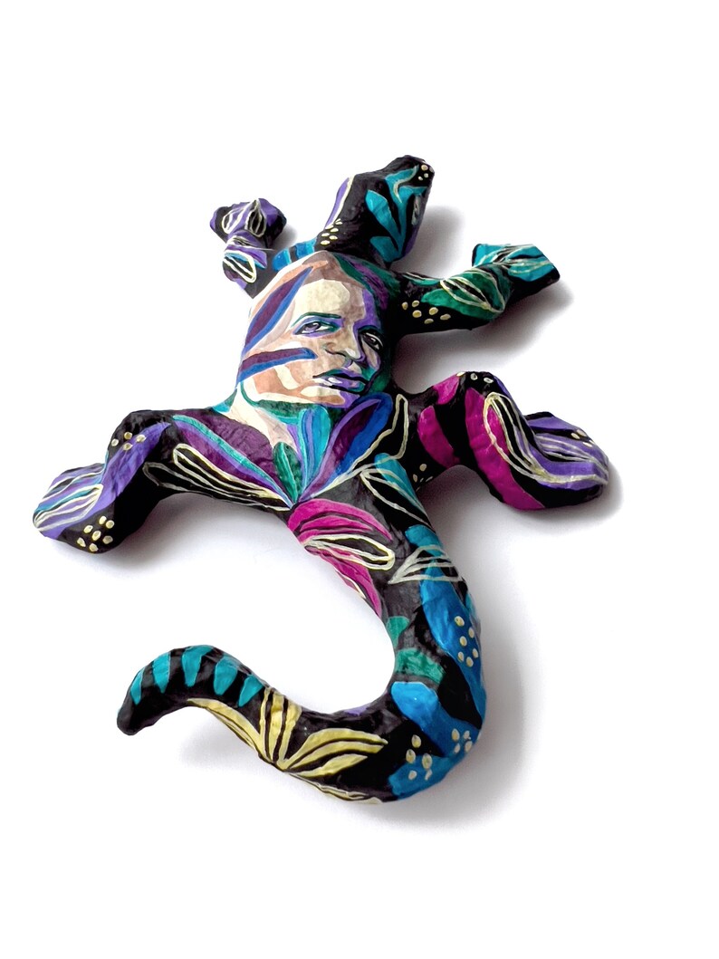 May include: A colorful, hand-painted paper mache lizard sculpture with a human face. The lizard is painted in shades of black, blue, green, purple, and pink with gold accents. The face is painted with realistic features and has a thoughtful expression.