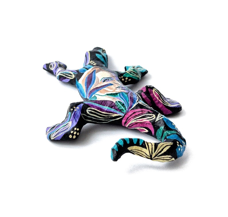 May include: A black painted gecko figurine with a colorful floral design. The gecko is lying on its belly with its tail curled up.