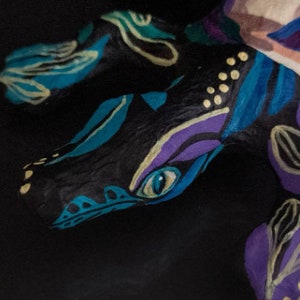 May include: A hand-painted ceramic sculpture of a dragon with blue, purple, and yellow accents. The dragon's eye is painted a bright blue.
