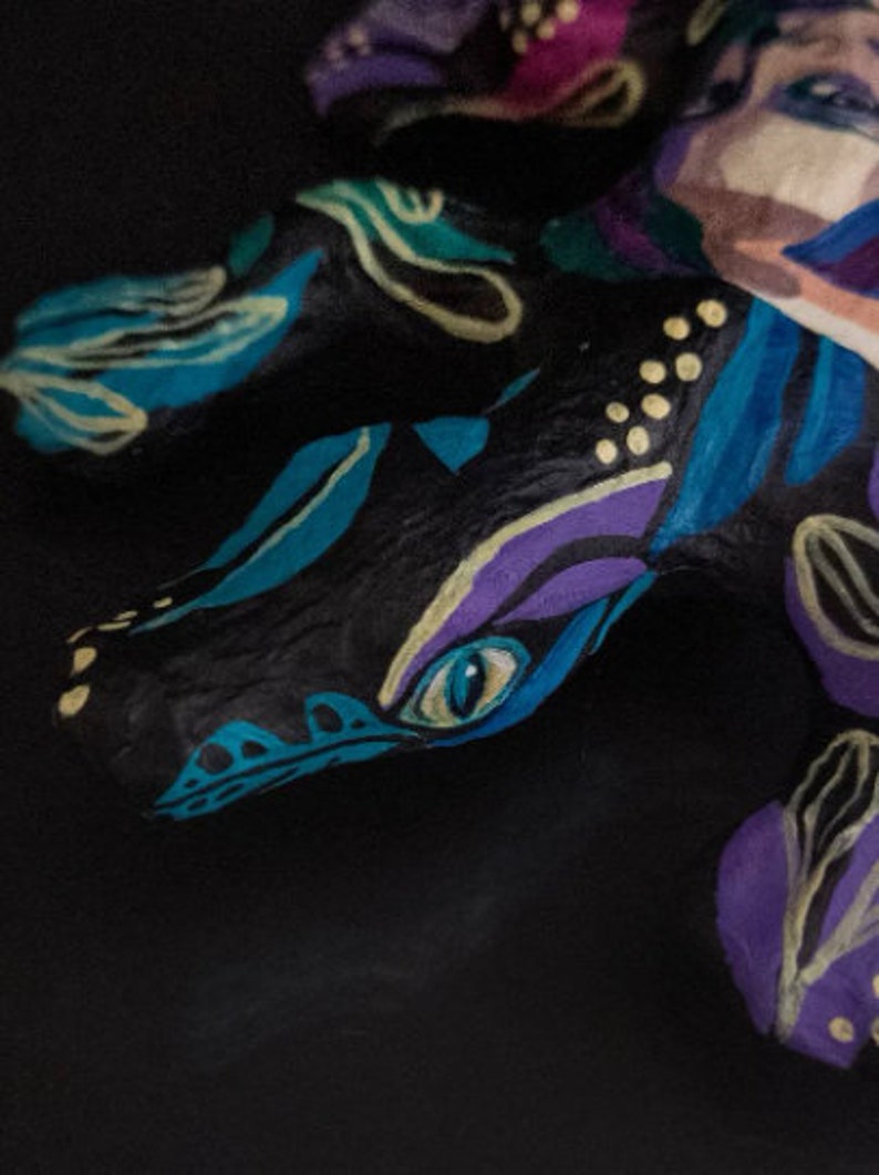 May include: A hand-painted ceramic sculpture of a dragon with blue, purple, and yellow accents. The dragon's eye is painted a bright blue.
