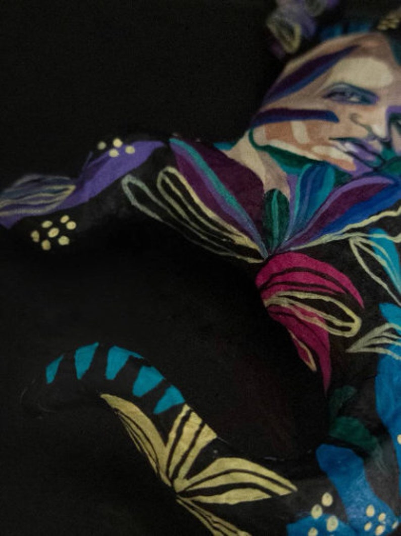 May include: A person with their body painted in a colorful floral pattern. The pattern includes shades of blue, purple, pink, yellow, and green. The person's face is also painted with a floral design.