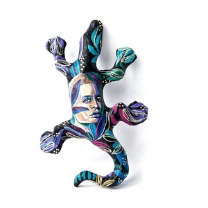 May include: A black paper mache lizard sculpture with a painted human face and colorful floral patterns. The lizard is in a curled up position with its tail curled around its body.